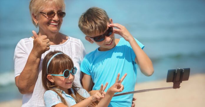Eine Oma posiert mit ihrem Enkel und ihrer Enkelin vor der Kamera, die der Enkel auf einem Selfie-Stick vor sich hinhält.