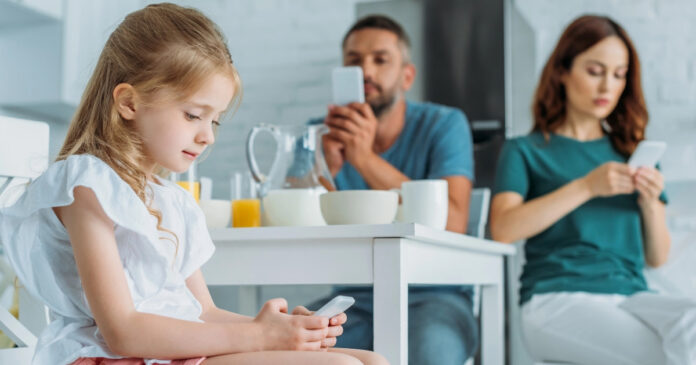 Kind und Eltern sitzen am Esstisch mit Essen auf dem Tisch. Alle drei sind am Smartphone