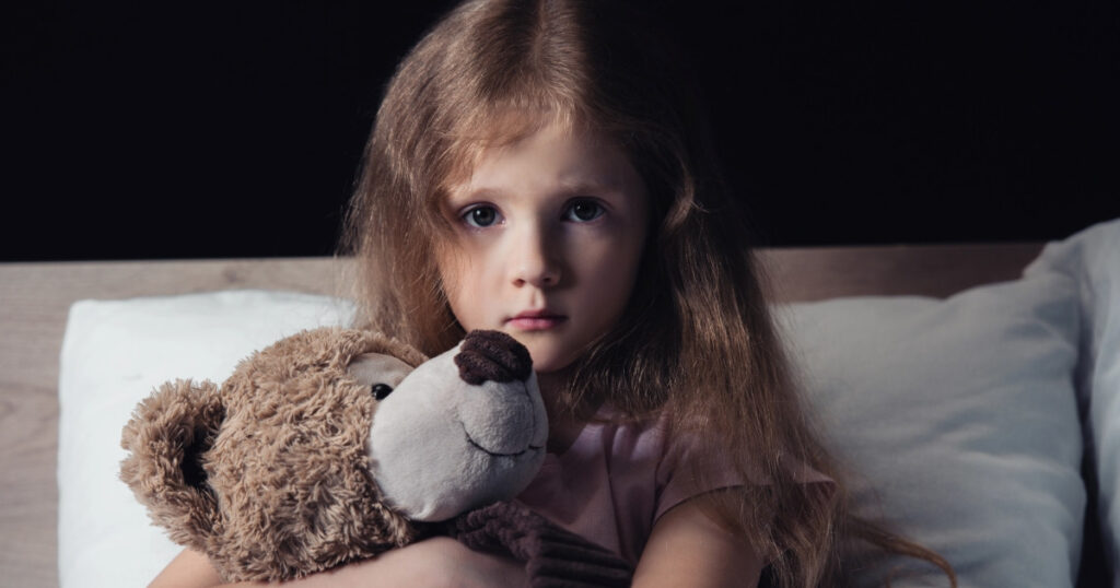 Ein Kind sitzt mit Teddy im Arm auf einem Bett in einem dunklen Raum
