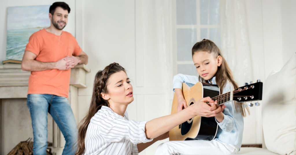 Eine Mutter hilft ihrer Tochter beim Gitarrenspiel. Der Vater schaut im Hintergrund zu