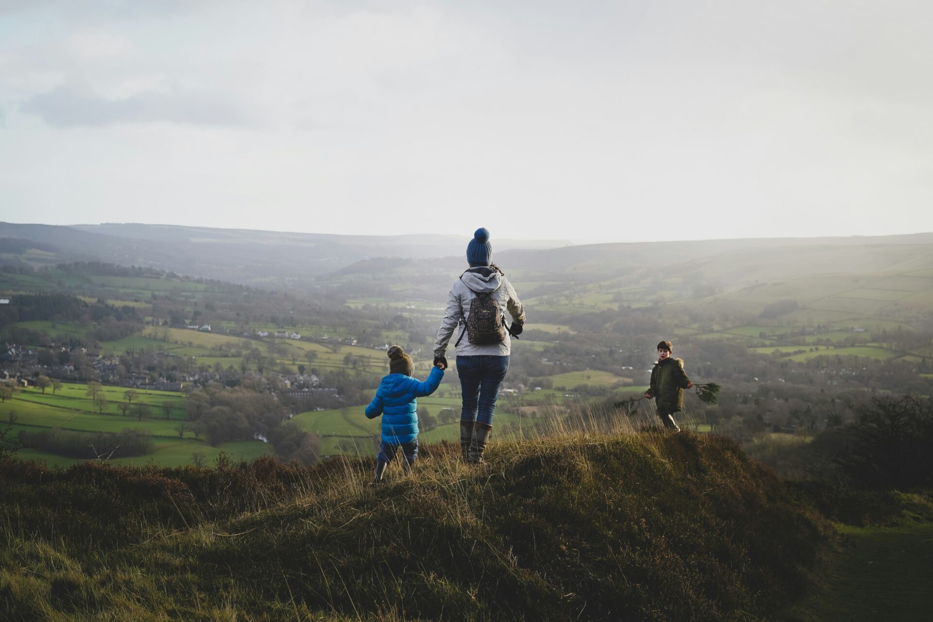 Elternteil geht mit zwei Kindern durch eine hügelige Landschaft und hält ein Kind an der Hand.