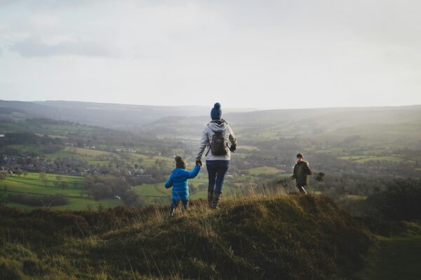 Elternteil geht mit zwei Kindern durch eine hügelige Landschaft und hält ein Kind an der Hand.
