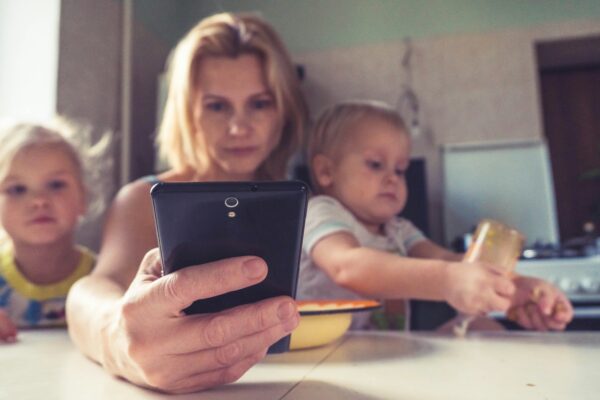 Mutter schaut auf ihr Smartphone, während ihre Kinder am Tisch sitzen.