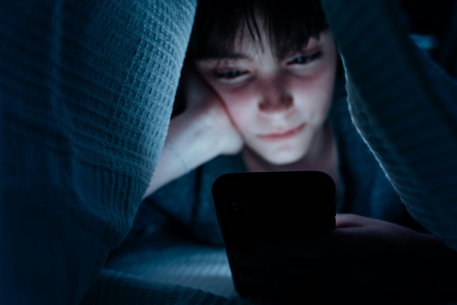 Jugendliche Person schaut abends unter einer Decke auf ihr Smartphone