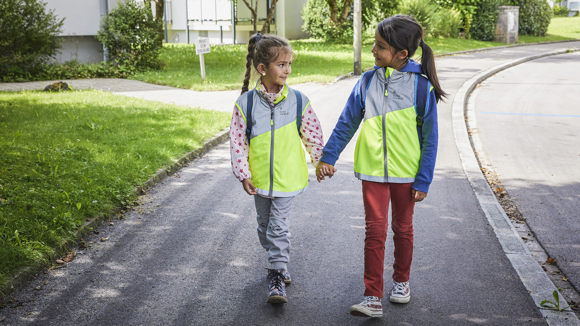 Zwei Kinder mit Leuchtwesten gehen auf einem Trottoir